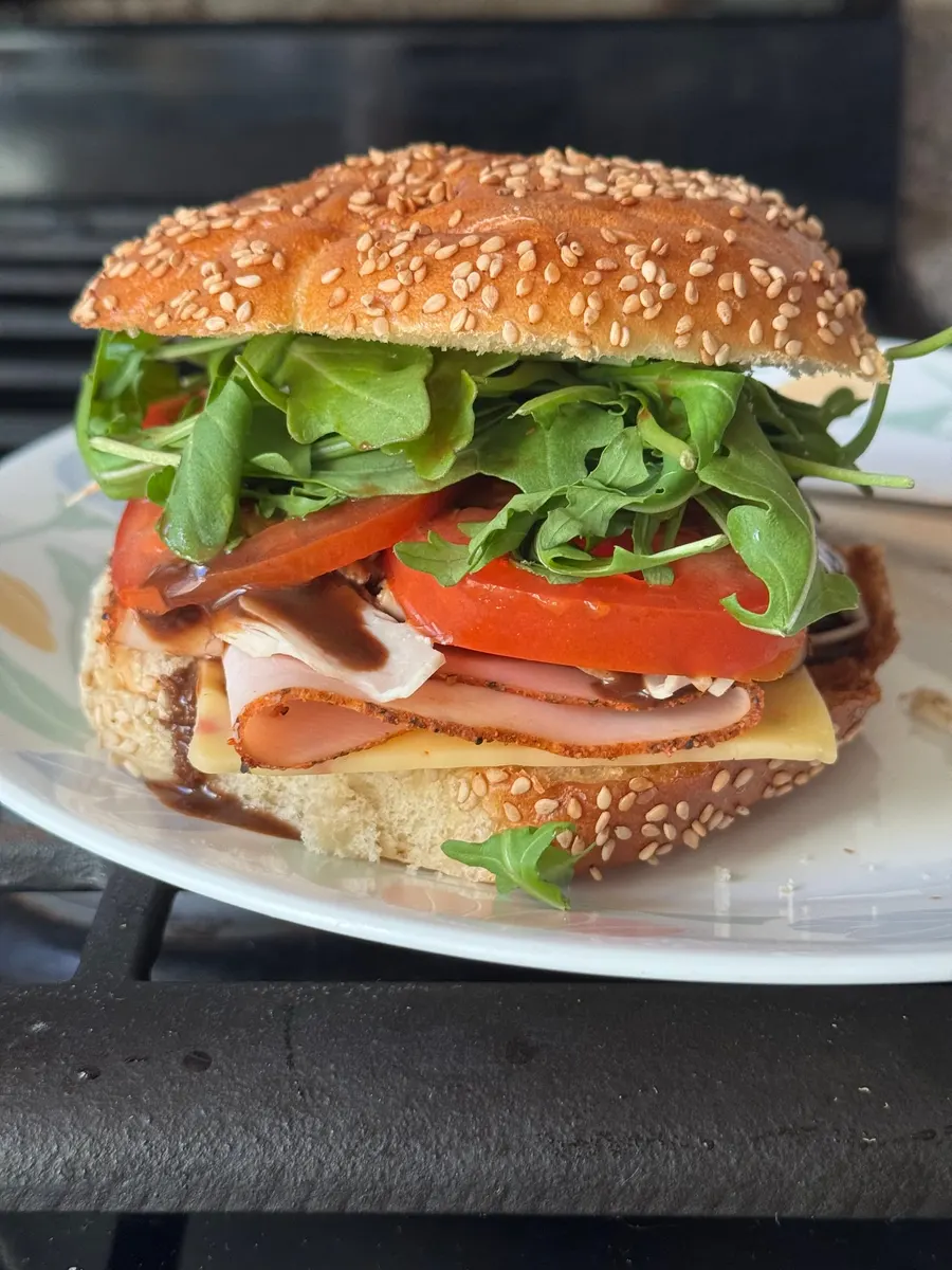 Tall sesame-roll sandwich layered with sliced deli meat, cheese, tomato, arugula, and dark sauce on a white plate.